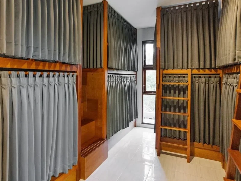 Dormitory room interior at In Da Surf Bali showing wooden display stands with hanging fabric curtain samples in various colors.