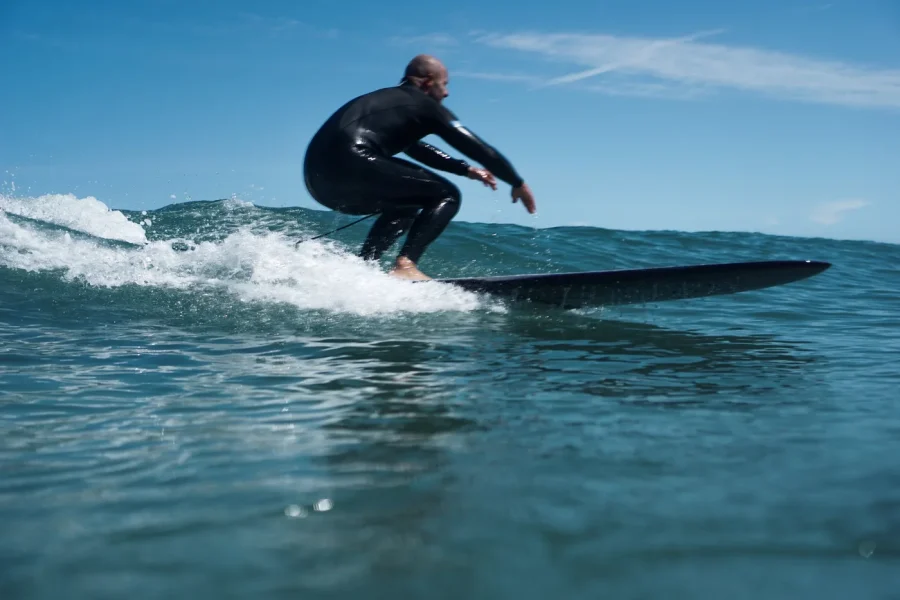 A surfer in a black wetsuit rides a small wave near Bidart, France with clear skies and calm ocean conditions visible.