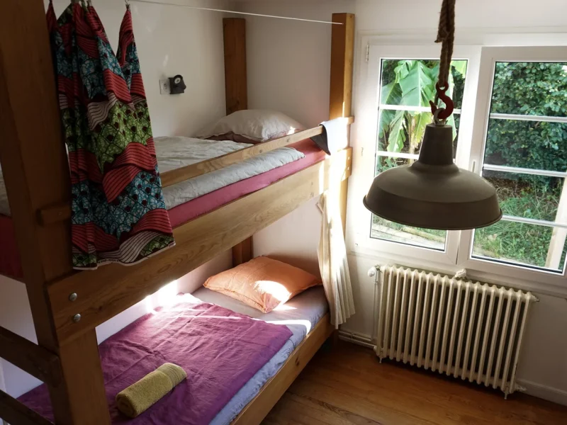 Shared room at Joyu Surf House in Bidart with wooden bunk beds, tropical fabrics, and views to banana plants outside.