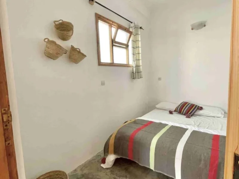 Chambre double à N'tona Surf Camp à Imsouane avec paniers tissés au mur, cadre de fenêtre en bois et couvre-lit rayé.