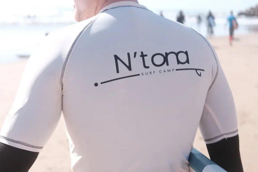Person mit weißem N'tona-Surf-Camp-Rashguard am Strand von Imsouane mit anderen Strandbesuchern im Hintergrund.