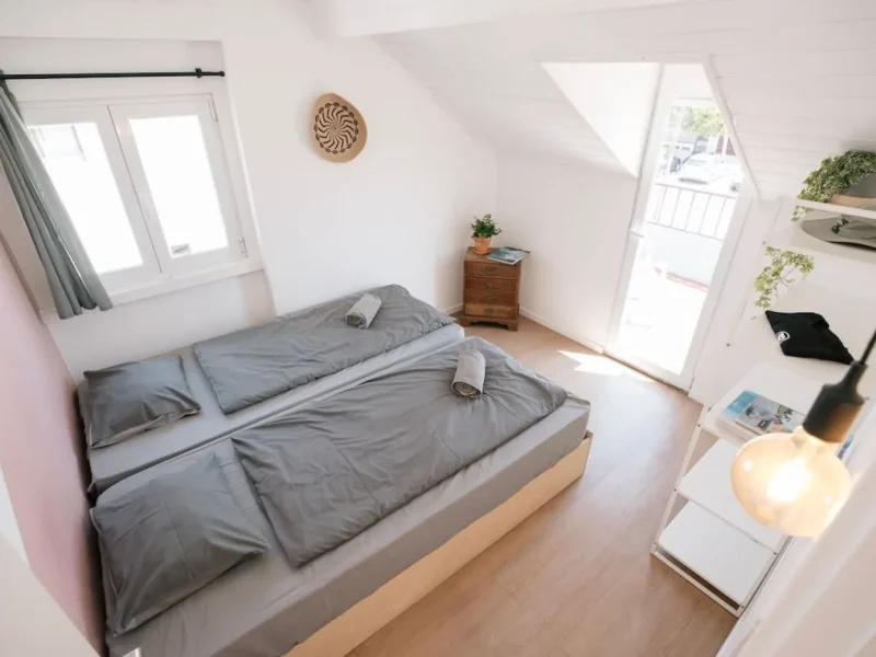 Zwei Einzelbetten mit grauer Bettwäsche in einem hellen Dachgeschoss-Schlafzimmer mit Dachfenstern und Holznachttisch im Surfbase Lisbon.