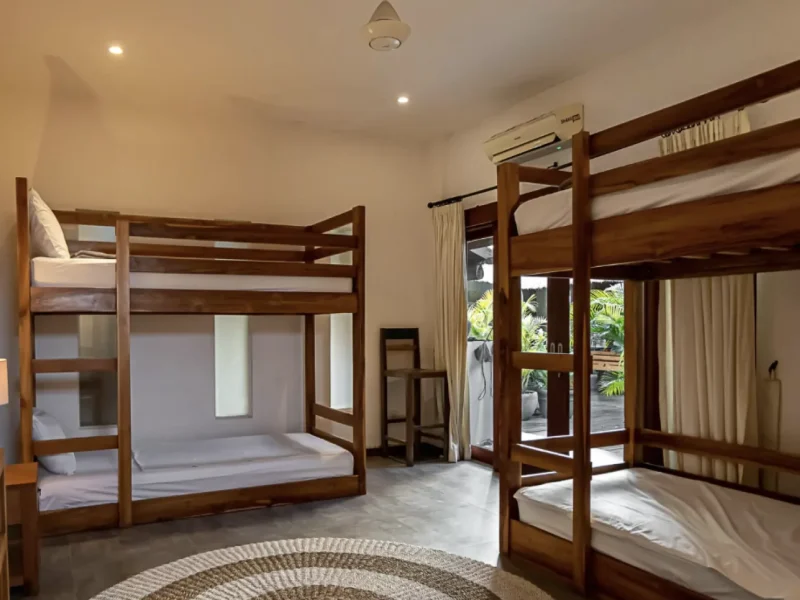 Mixed shared dormitory room with wooden bunk beds, air conditioning, and glass doors opening to tropical garden views.