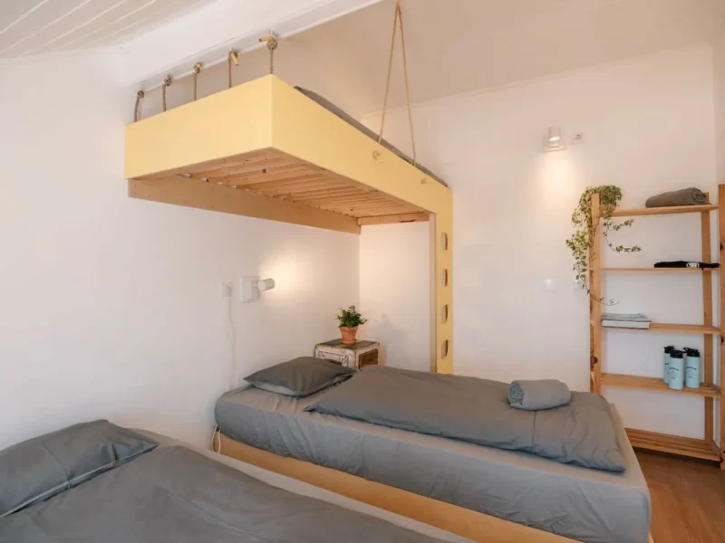 Dreibettzimmer im Surfbase Lisbon mit einem hängenden gelb-hölzernen Bettgestell, grauer Bettwäsche und Topfpflanzen.