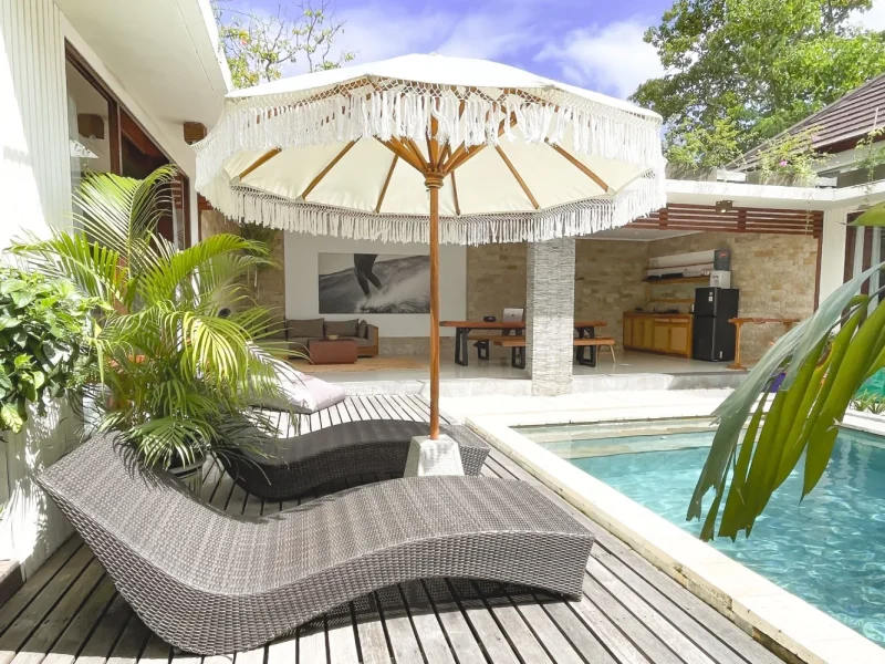 Outdoor terrace with swimming pool, loungers and white fringe umbrella overlooking accommodation at CARI Surf Camp in Uluwatu, Bali.