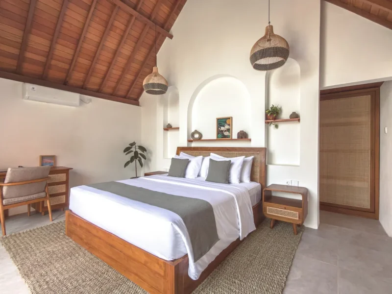 Private bedroom at CARI Surf Camp in Uluwatu featuring wooden bed frame with woven rattan headboard, white bedding, and arched alcove wall.