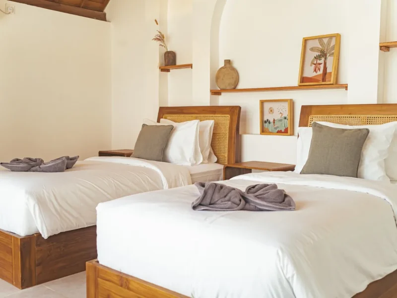 Mixed shared twin room at CARI Surf Camp in Uluwatu features wooden beds with rattan headboards, white linens, and wooden wall shelving with decorative pottery.