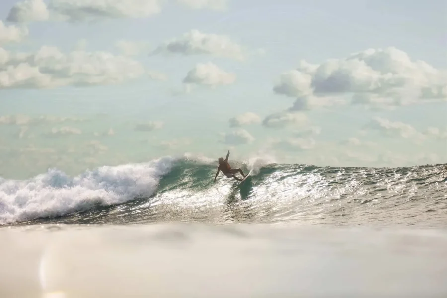 Surfer riding a wave at Uluwatu, Bali with sandy beach and cloudy sky.