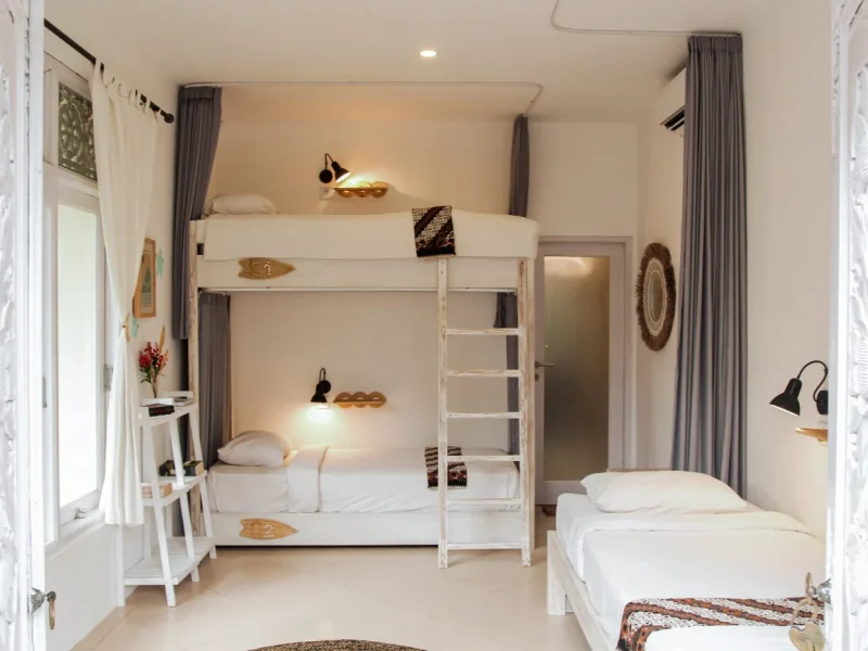 A bright female shared room at Mondo Surf Village features a white bunk bed with individual reading lights and a single bed with ocean-themed decor accents.