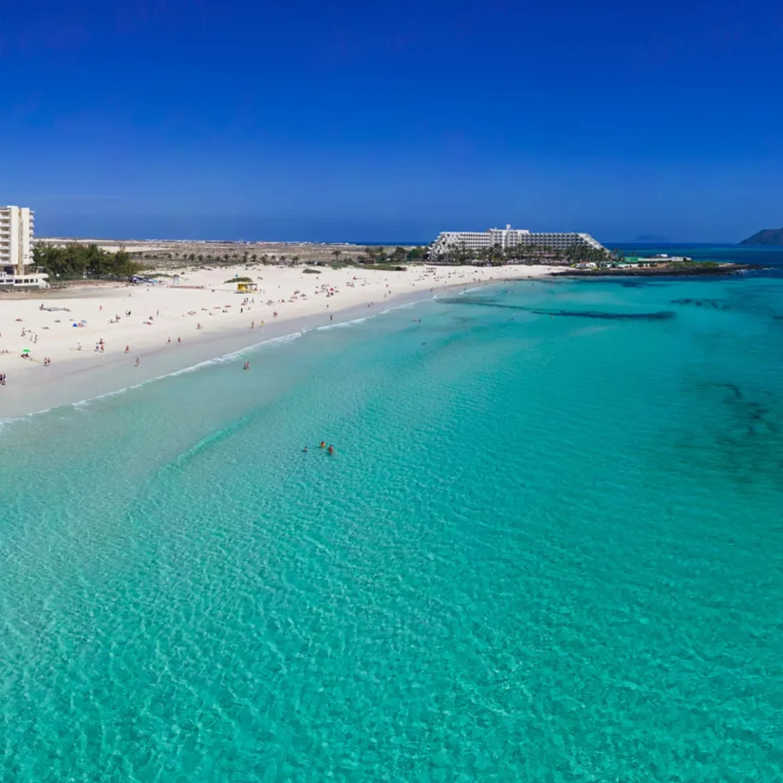 Plage immaculée à Fuerteventura avec eau turquoise cristalline, sable blanc, bâtiments du complexe et baigneurs le long du rivage.