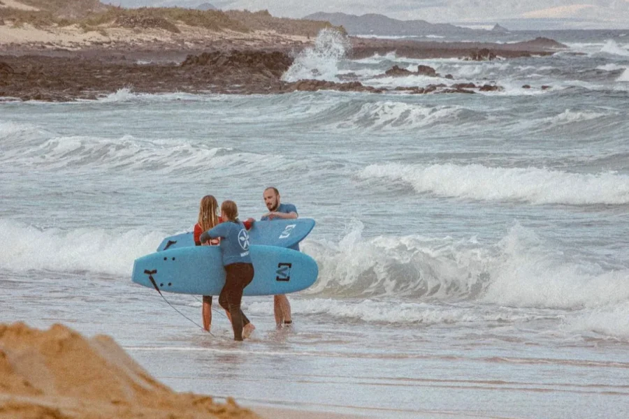 Erwachsener Instruktor und zwei Kinder halten blaue Surfbretter im flachen Wasser am Strand von Fuerteventura.