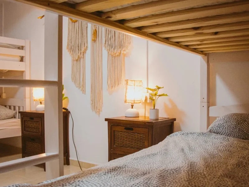 Gemischtes Schlafsaal-Schlafzimmer mit Holzbalken, Makramee-Wandbehängen und geflochtenen Nachttischmöbeln in der International Surf School & Camp Fuerteventura.