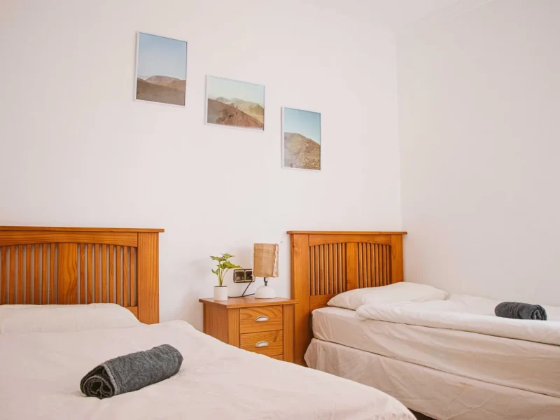 Zweibettzimmer mit Holz-Kopfteilen, weißer Bettwäsche und drei gerahmten Wüstenlandschaftsfotos an weißer Wand.