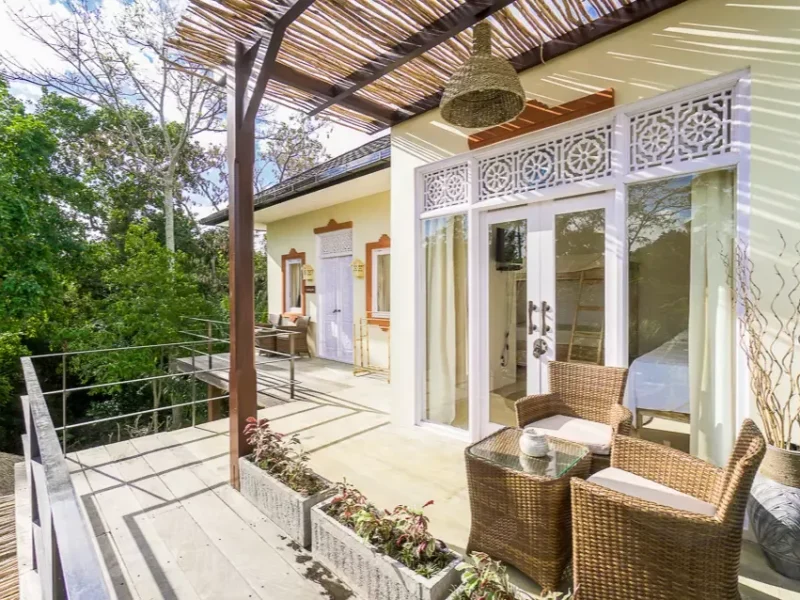 The terrace of a shared accommodation room features wicker seating, pergola shade structure, and white railing overlooking tropical vegetation in Canggu.
