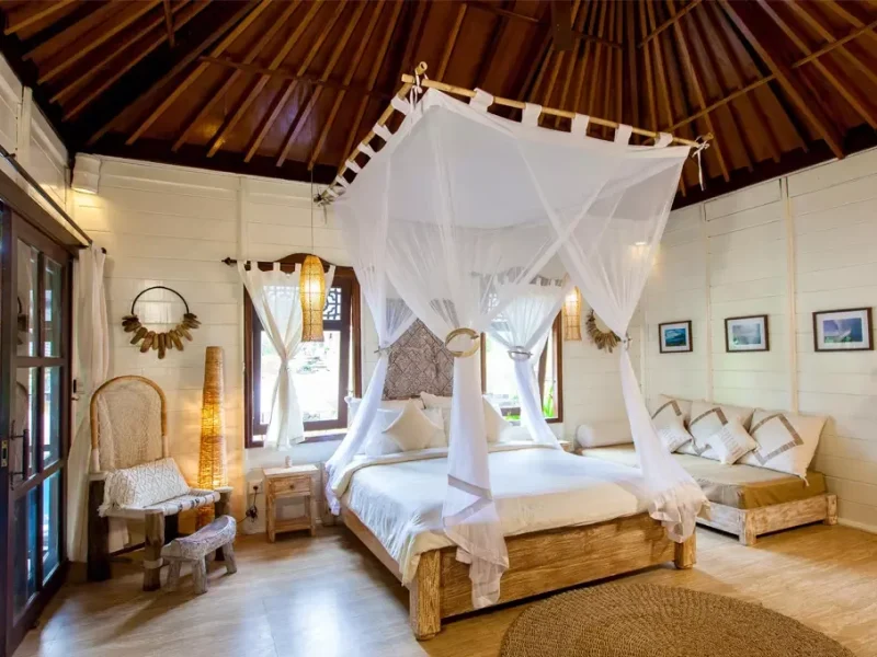Private bungalow bedroom at Mondo Surf Village featuring a four-poster canopy bed with white drapes and wooden ceiling beams.