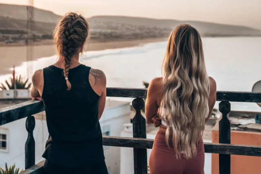 Two women stand on a terrace railing overlooking the Taghazout coastline and beach at sunset.
