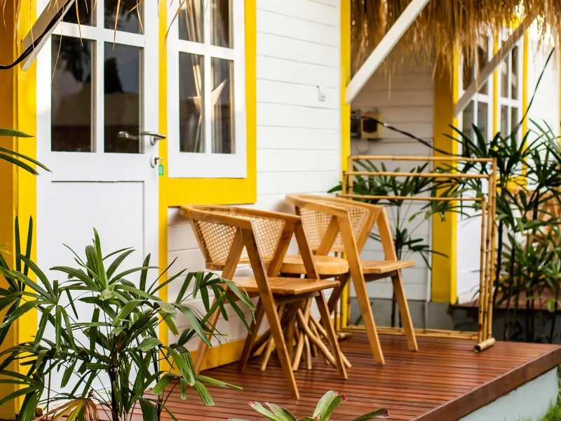 Wooden cabin terrace at Soleia Surf Canggu featuring yellow-trimmed architecture, woven rattan chairs, and potted tropical plants.