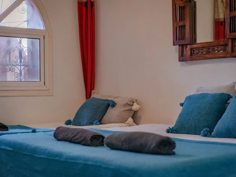 Privatzimmer im The Little Kasbah mit Twin-Betten, blauer Bettwäsche, gerollten Handtüchern und traditioneller hölzerner Wanddekoration in Imsouane.