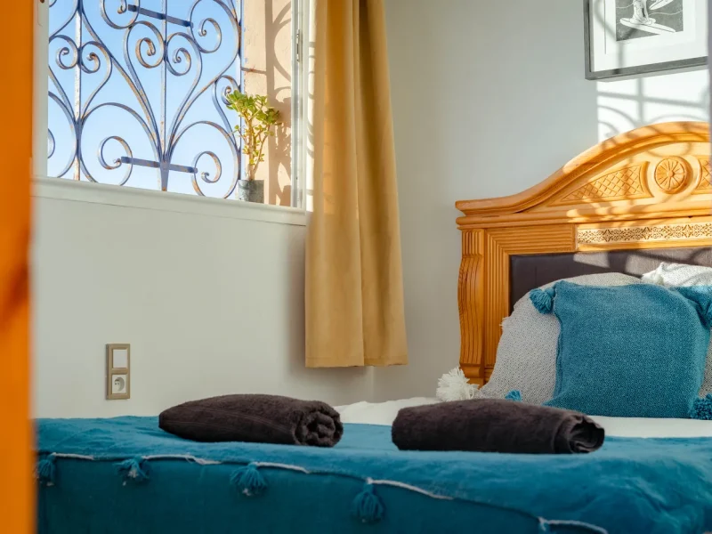 Privatzimmer im The Little Kasbah mit hölzernem Bettrahmen mit dekorativem Kopfteil, türkisfarbener Bettwäsche, gerollten Handtüchern und kunstvollem Fenster mit Eisengitter mit Blick auf Imsouane.