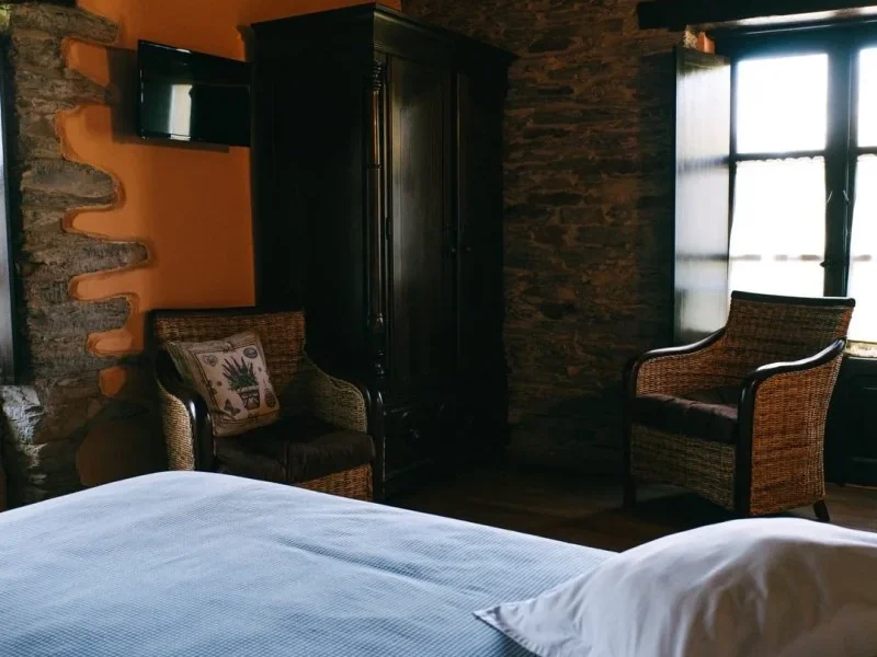 Private double bedroom featuring exposed stone walls, blue bedding, wicker chairs, and contemporary orange wall art at Enjoy Native in Asturias.