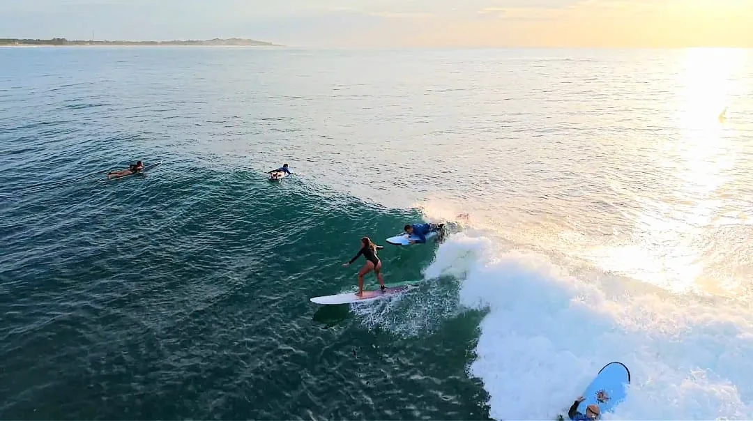 Tres surfistas surfeando y remando olas en Arugam Bay al atardecer con la costa de Wanderlust Surf Camp, Sri Lanka