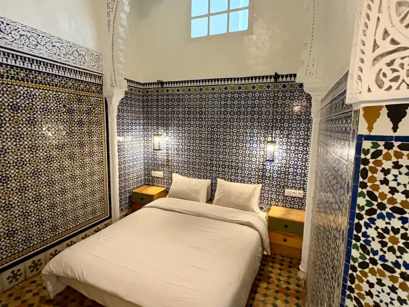 Double bed in a room at Ilyana Surf House Rabat, surrounded by traditional Moroccan zellige tile patterns and carved plaster details.