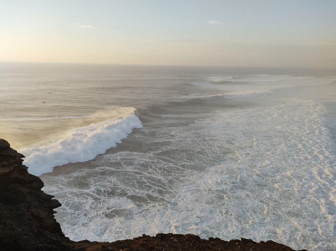 Ein Surfer paddelt bei Sonnenuntergang an Praia do Norte in Nazaré, Portugal, mit warmem goldenem Licht über dem Atlantik und sauberen Wellengesichtern dahinter.