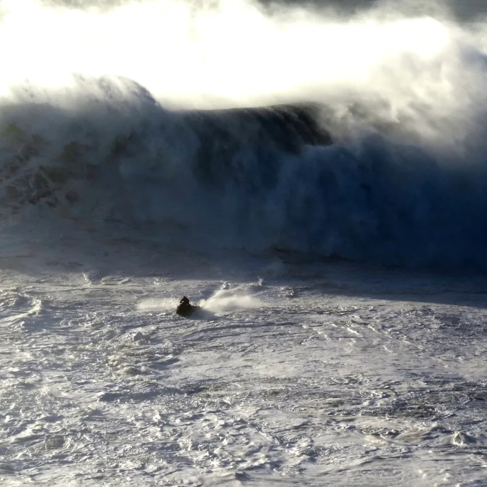 A surfer being towed into a giant wave at Praia do Norte in Nazaré, with white spray flying off the crest and the underwater Nazaré canyon producing the dramatic vertical wall.