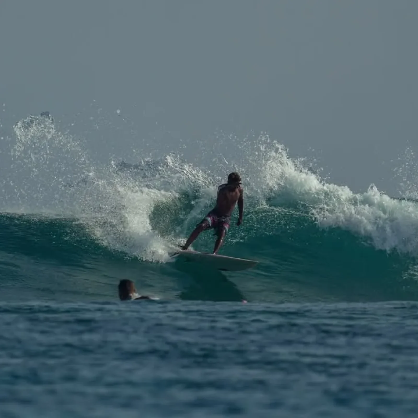 Surfer on board riding a breaking wave at Kudahuvadhoo with another person swimming nearby.