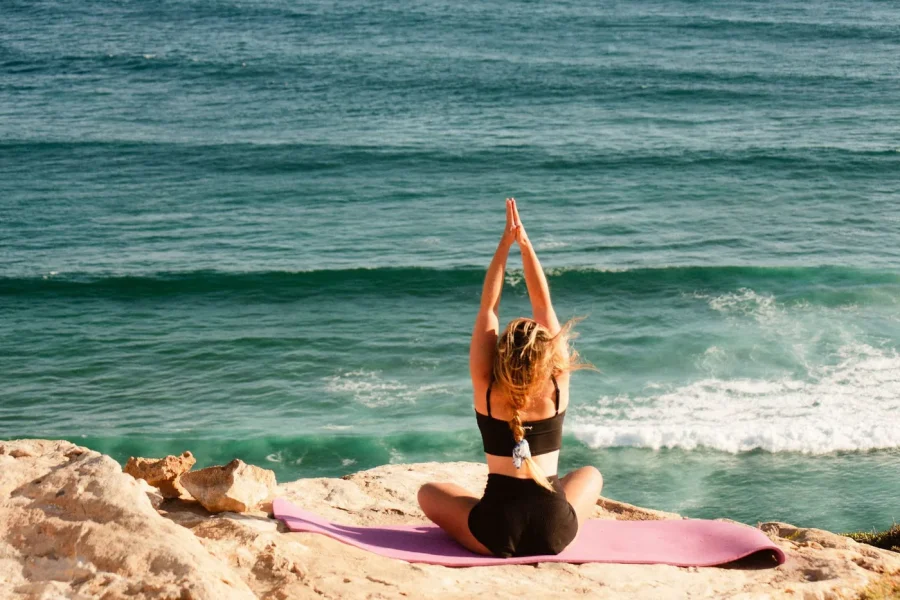 Eine Frau führt eine Yogapose auf einer rosa Matte am Strand von Imsouane aus, im Hintergrund sind Wellen sichtbar.