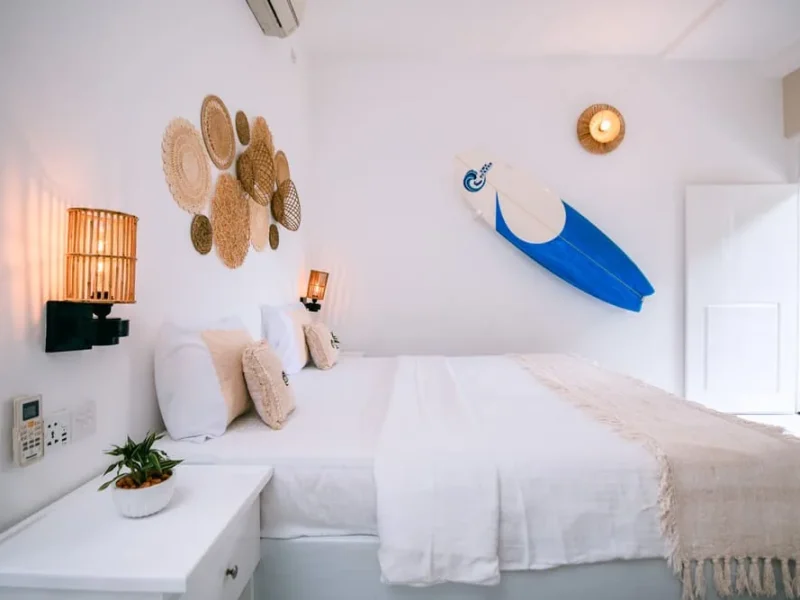 Private single bedroom with white bedding, woven plate wall art, blue surfboard decoration, and wicker furniture.