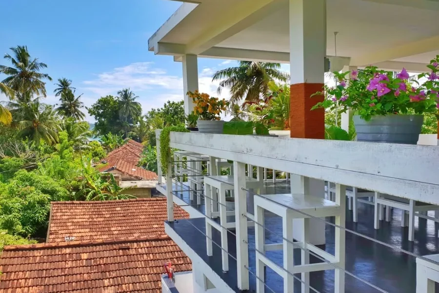 TS2 Weligama Surf Camp's upper level terrace features white architectural railings, potted flowering plants, and views over the surrounding Weligama area.