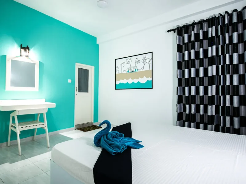 Private double/twin room interior at TS2 Weligama Surf Camp featuring turquoise accent wall, white bed with blue towel, and coastal-themed artwork.