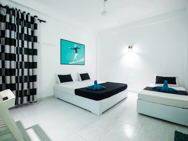 Private triple room at TS2 Weligama Surf Camp features white platform beds with black bedding and surf-inspired wall art depicting a surfer on turquoise background.