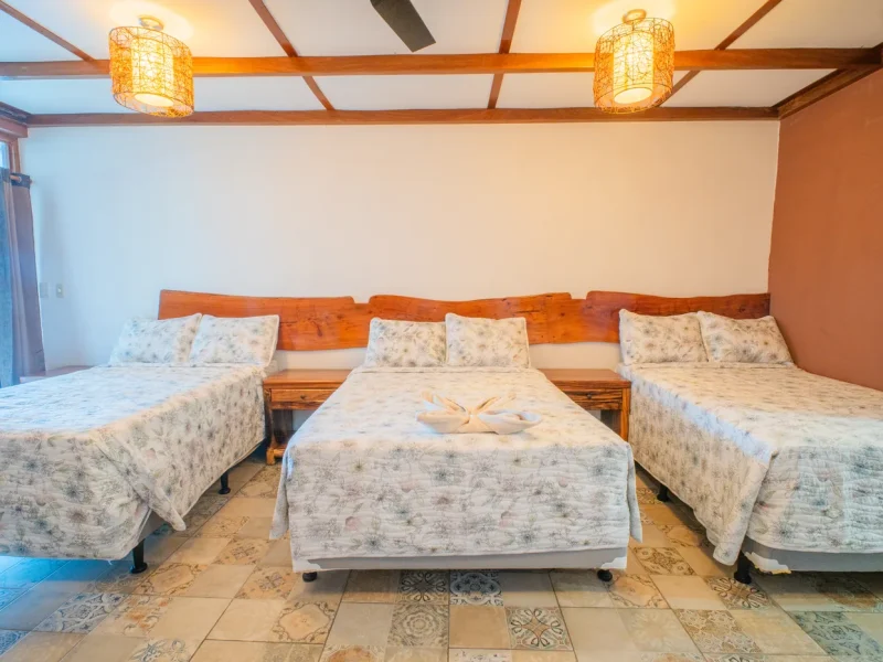 Dormitorio de la Triple Poolside Suite con tres camas, vigas de cabecero de madera, lámparas colgantes trenzadas y suelo de baldosas decorativas.