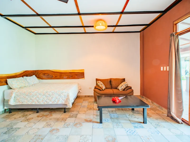 Dormitorio de la Single Poolside Suite en Casa Las Flores con cama con cabecero de madera, paredes naranjas, vigas decorativas en el techo y zona de asientos con suelo de baldosas estampadas.