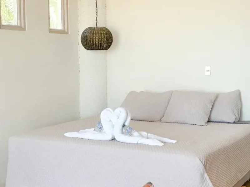 Single suite bedroom at Punta Mango Surf Resort with bed, decorative towel sculpture, woven pendant light, and beige furnishings.