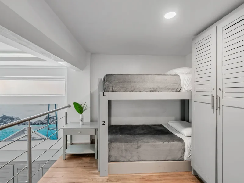 Dorm room at Iguana Surf Boutique Hotel in Tamarindo with two white bunk beds, grey bedding, and partial ocean view through window.