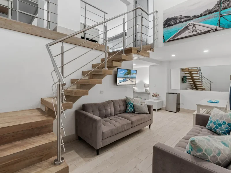 Family Suite interior with floating wooden staircase, gray upholstered sofa, and beach-themed wall art in Tamarindo.