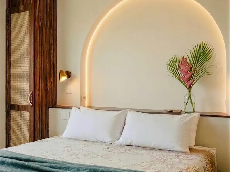Modern hotel room featuring a king bed with white pillows and a striking illuminated arched neon headboard in warm white light, with a fresh pink flower arrangement on the bedside shelf.