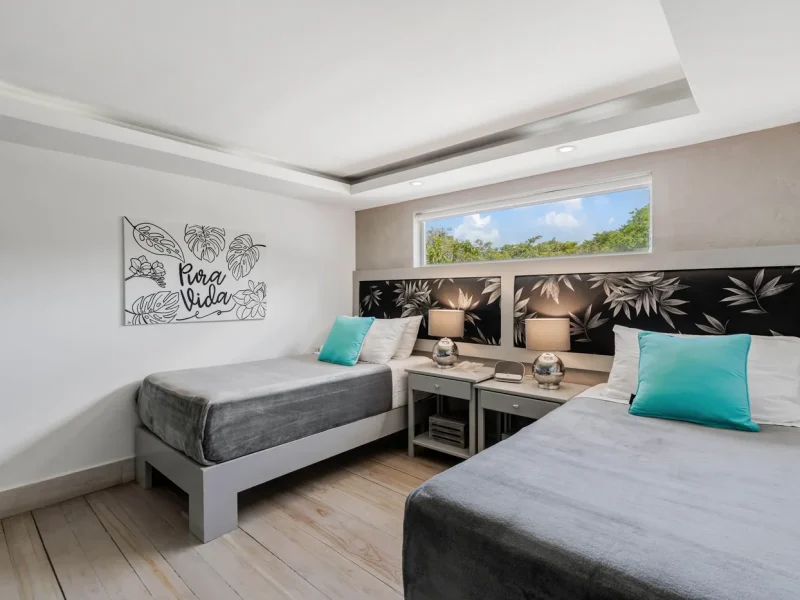 Private twin bedroom at Iguana Surf Boutique Hotel featuring two single beds with gray linens, turquoise pillows, and botanical wall art.