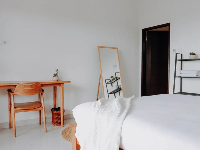 A minimalist bedroom at Elsewhere Surf Camp Bali in Canggu features an orange wood bed frame, matching desk and chair, standing mirror, and shelving unit.